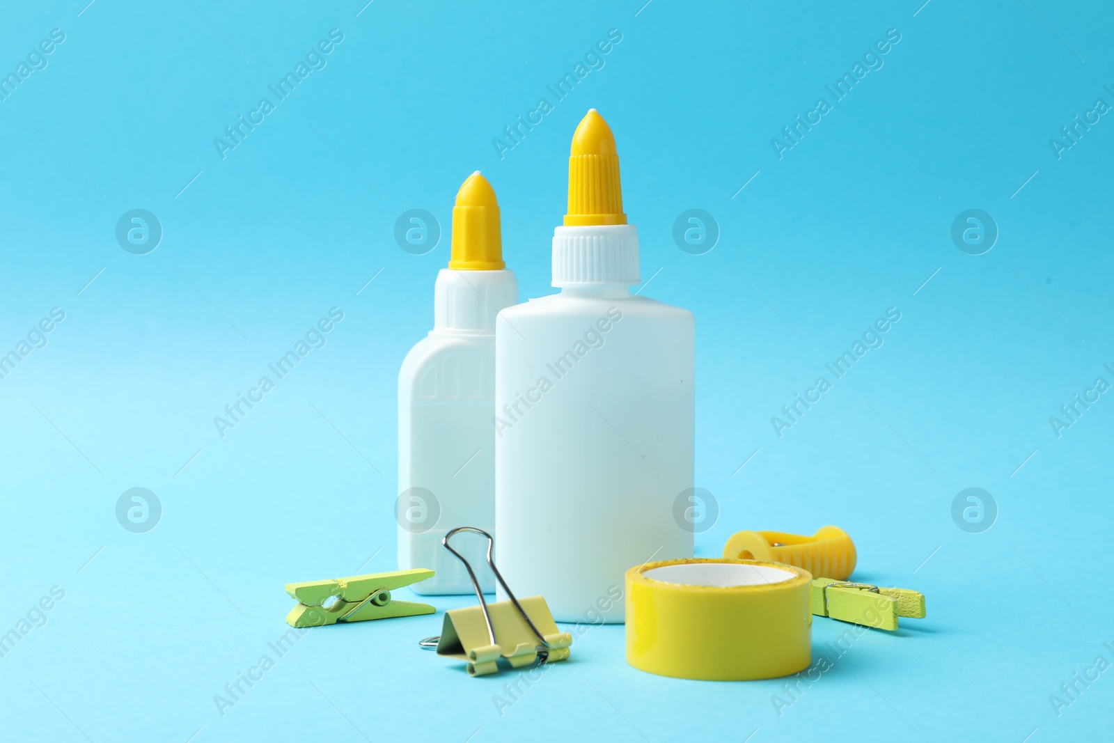 Photo of Glue in bottles, binder clip, sharpener and adhesive tape on light blue background, closeup