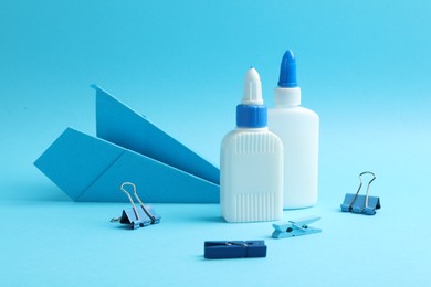Photo of Glue in bottles, binder clips and paper plane on light blue background, closeup