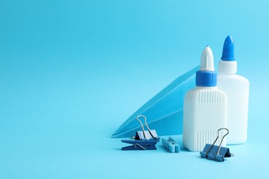 Photo of Glue in bottles, binder clips and paper plane on light blue background, closeup. Space for text