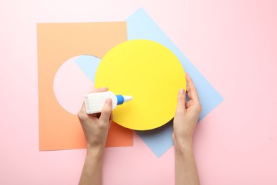 Photo of Woman making craft with glue and color paper at pink table, top view