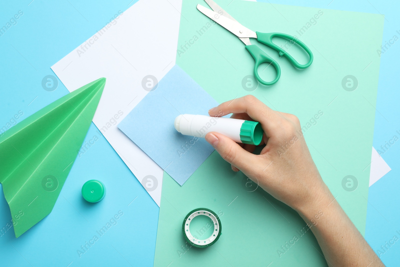 Photo of Woman making craft with glue and color paper at light blue table, top view