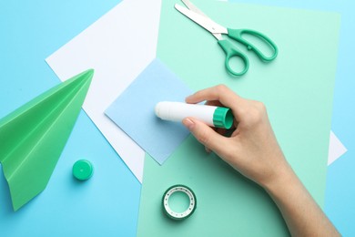 Photo of Woman making craft with glue and color paper at light blue table, top view