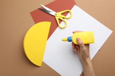 Photo of Woman making craft with glue and color paper at brown table, top view