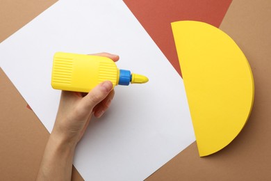Photo of Woman making craft with glue and color paper at brown table, top view