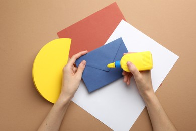 Photo of Woman making craft with glue and color paper at brown table, top view