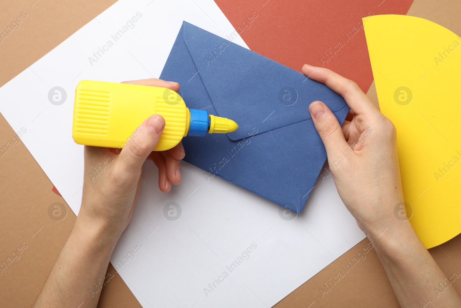 Photo of Woman making craft with glue and color paper at brown table, top view