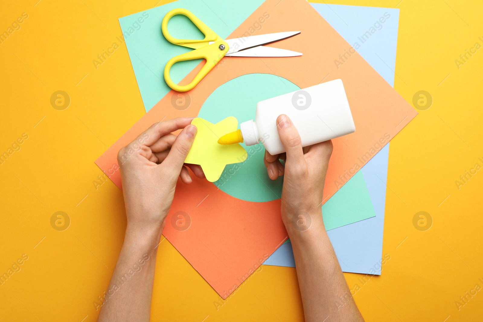 Photo of Woman making craft with glue and color paper at orange table, top view