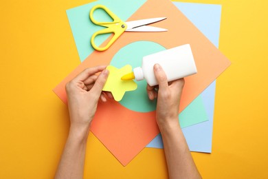Photo of Woman making craft with glue and color paper at orange table, top view