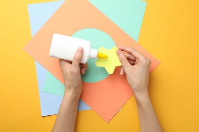 Photo of Woman making craft with glue and color paper at orange table, top view