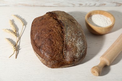 Photo of Loaf of fresh bread, spikelets, flour and rolling pin on white wooden table