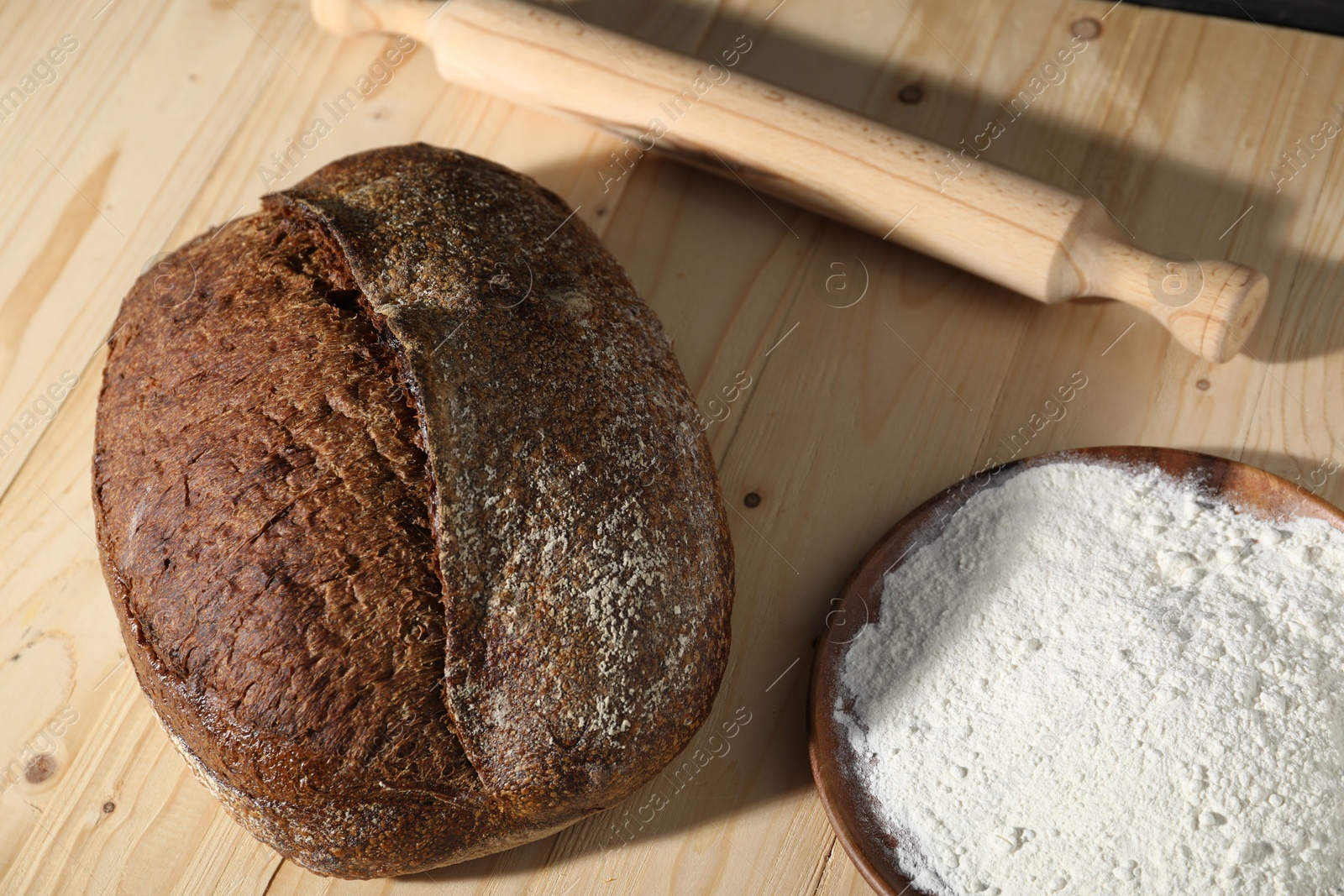 Photo of Loaf of fresh bread, flour and rolling pin on light wooden table, above view