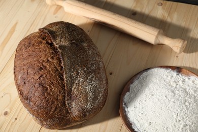 Photo of Loaf of fresh bread, flour and rolling pin on light wooden table, above view