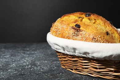 Photo of Loaf of fresh bread with sesame seeds and raisins in wicker basket on grey textured table, closeup. Space for text