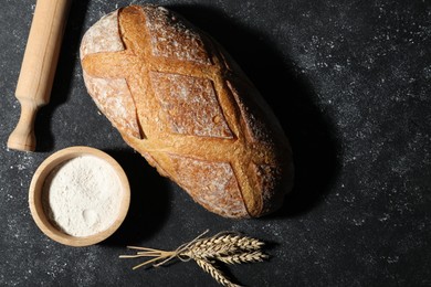 Photo of Loaf of fresh bread, rolling pin, flour and spikelets on grey textured table, flat lay. Space for text