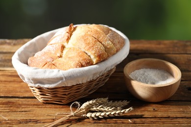 Photo of Fresh cut bread in wicker basket, flour and spikelets on wooden table against blurred background, closeup