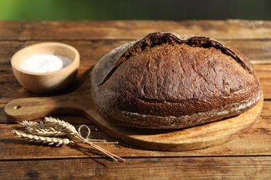 Photo of Loaf of fresh bread, flour and spikelets on wooden table against blurred background, closeup