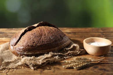 Photo of Loaf of fresh bread, flour and spikelets on wooden table against blurred background, closeup