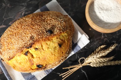 Photo of Loaf of fresh bread, flour and spikelets on black marble table, flat lay