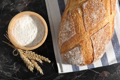 Photo of Loaf of fresh bread, flour and spikelets on black marble table, flat lay