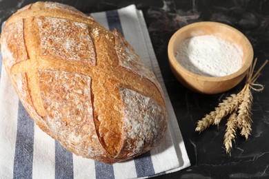 Photo of Loaf of fresh bread, flour and spikelets on black marble table, closeup