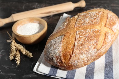 Photo of Loaf of fresh bread, flour, rolling pin and spikelets on black marble table, closeup