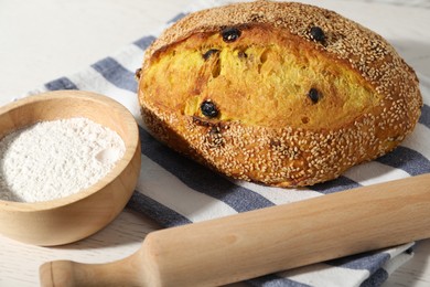 Photo of Loaf of fresh bread with sesame seeds and raisins, flour and rolling pin on white wooden table, closeup