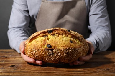 Photo of Baker with loaf of fresh bread with sesame seeds and raisins at wooden table, closeup