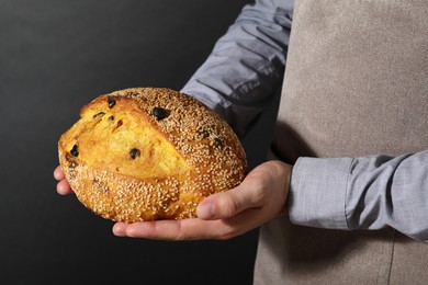 Photo of Baker with loaf of fresh bread with sesame seeds and raisins on black background, closeup