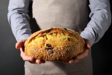 Photo of Baker with loaf of fresh bread with sesame seeds and raisins on black background, closeup