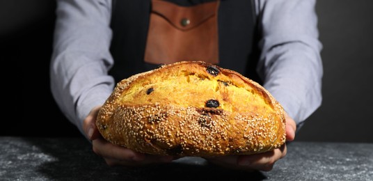 Photo of Baker with loaf of fresh bread with sesame seeds and raisins at grey textured table, closeup