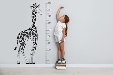 Image of Cute little girl standing on stacked books and measuring her height. White wall with scale and drawing of giraffe