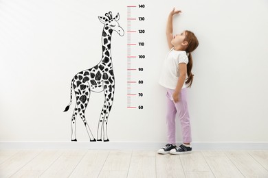 Image of Cute little girl measuring her height near white wall with scale and drawing of giraffe