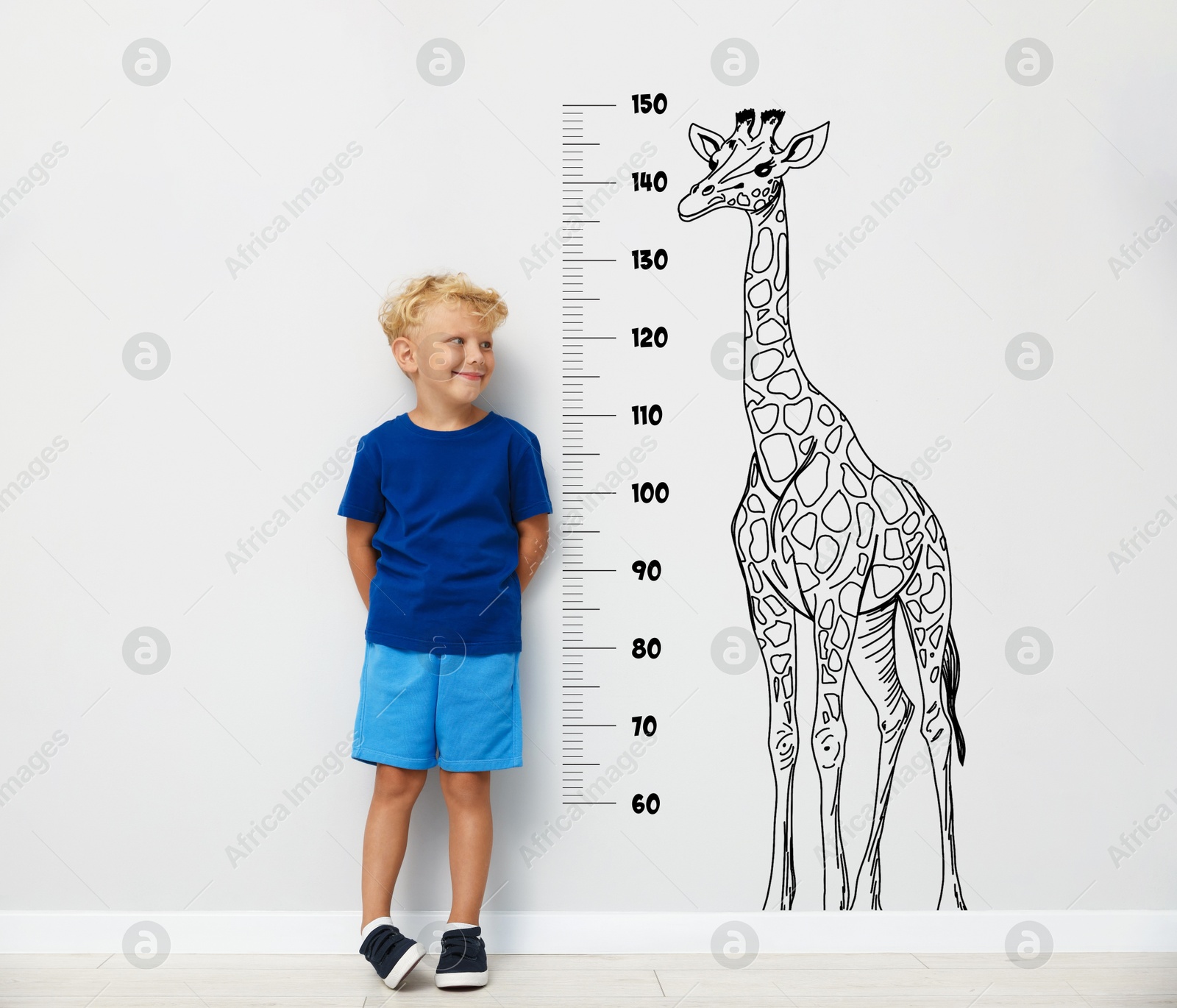 Image of Cute little boy measuring his height near white wall with scale and drawing of giraffe
