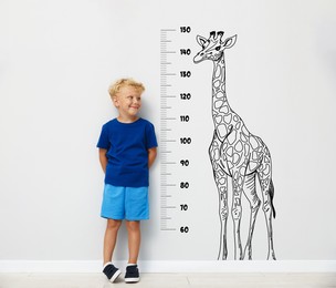 Image of Cute little boy measuring his height near white wall with scale and drawing of giraffe