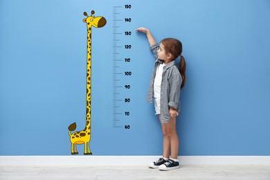 Image of Cute little girl measuring her height near blue wall with scale and drawing of giraffe