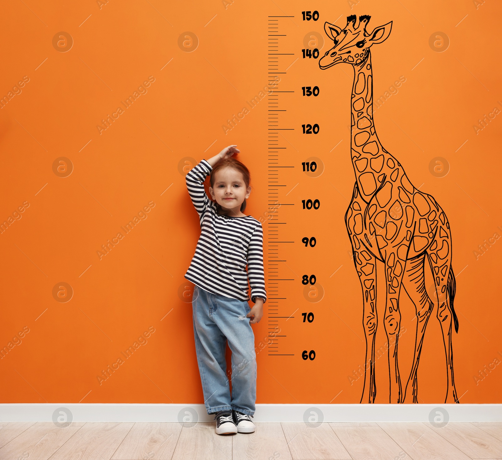Image of Cute little girl measuring her height near orange wall with scale and drawing of giraffe
