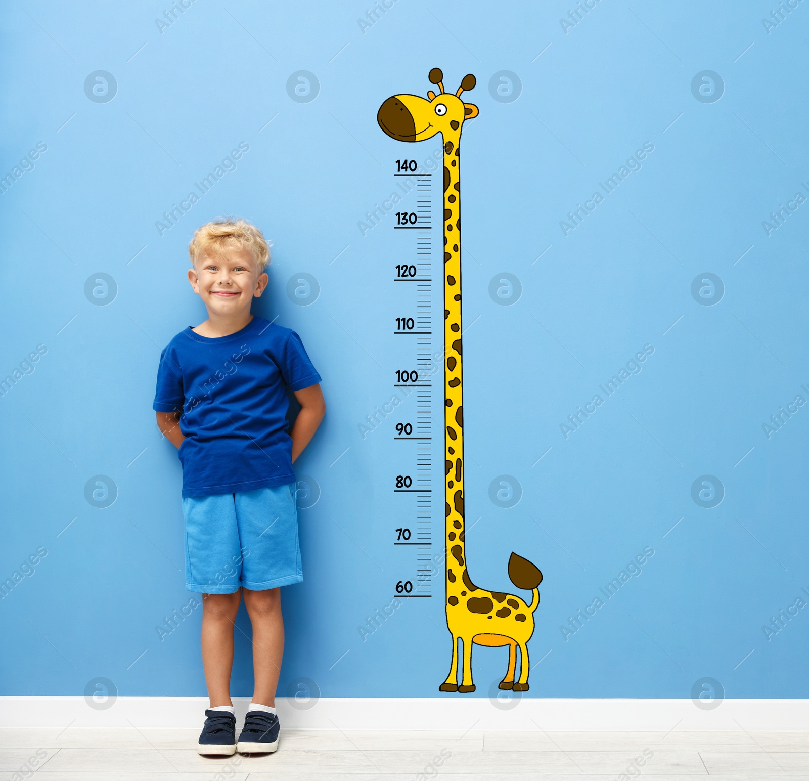 Image of Cute little boy measuring his height near blue wall with scale and drawing of giraffe