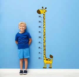 Image of Cute little boy measuring his height near blue wall with scale and drawing of giraffe