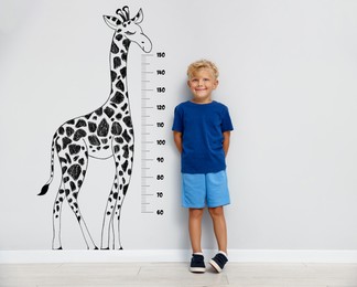 Image of Cute little boy measuring his height near white wall with scale and drawing of giraffe