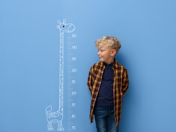 Image of Cute little boy measuring his height near blue wall with scale and drawing of giraffe