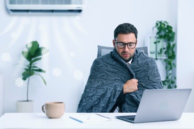 Image of Freezing worker wrapped in blanket at table in office. Broken or incorrectly adjusted air conditioner