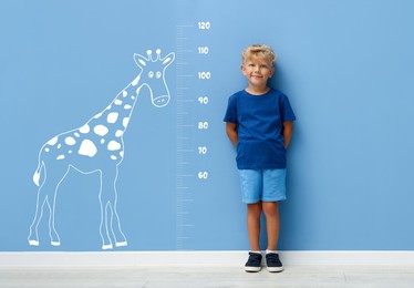 Image of Cute little boy measuring his height near blue wall with scale and drawing of giraffe
