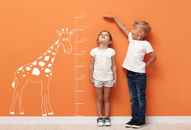 Image of Cute little children measuring their heights near orange wall with scale and drawing of giraffe