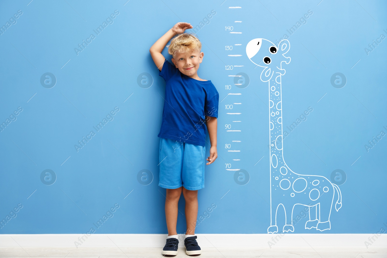 Image of Cute little boy measuring his height near blue wall with scale and drawing of giraffe