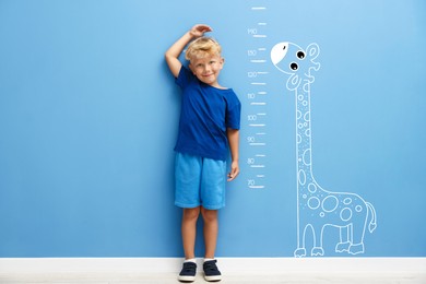 Image of Cute little boy measuring his height near blue wall with scale and drawing of giraffe