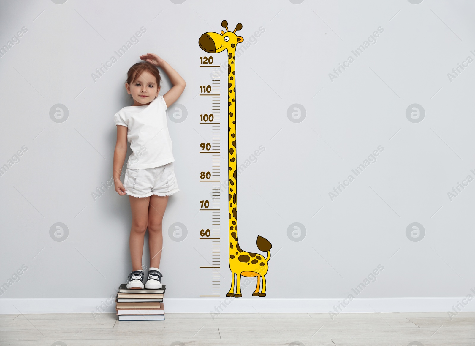 Image of Cute little girl standing on stacked books and measuring her height. White wall with scale and drawing of giraffe