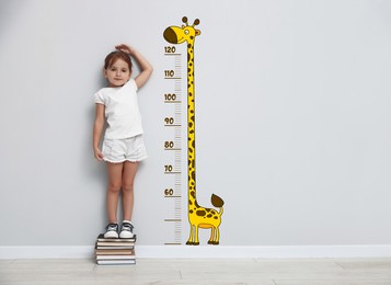 Image of Cute little girl standing on stacked books and measuring her height. White wall with scale and drawing of giraffe