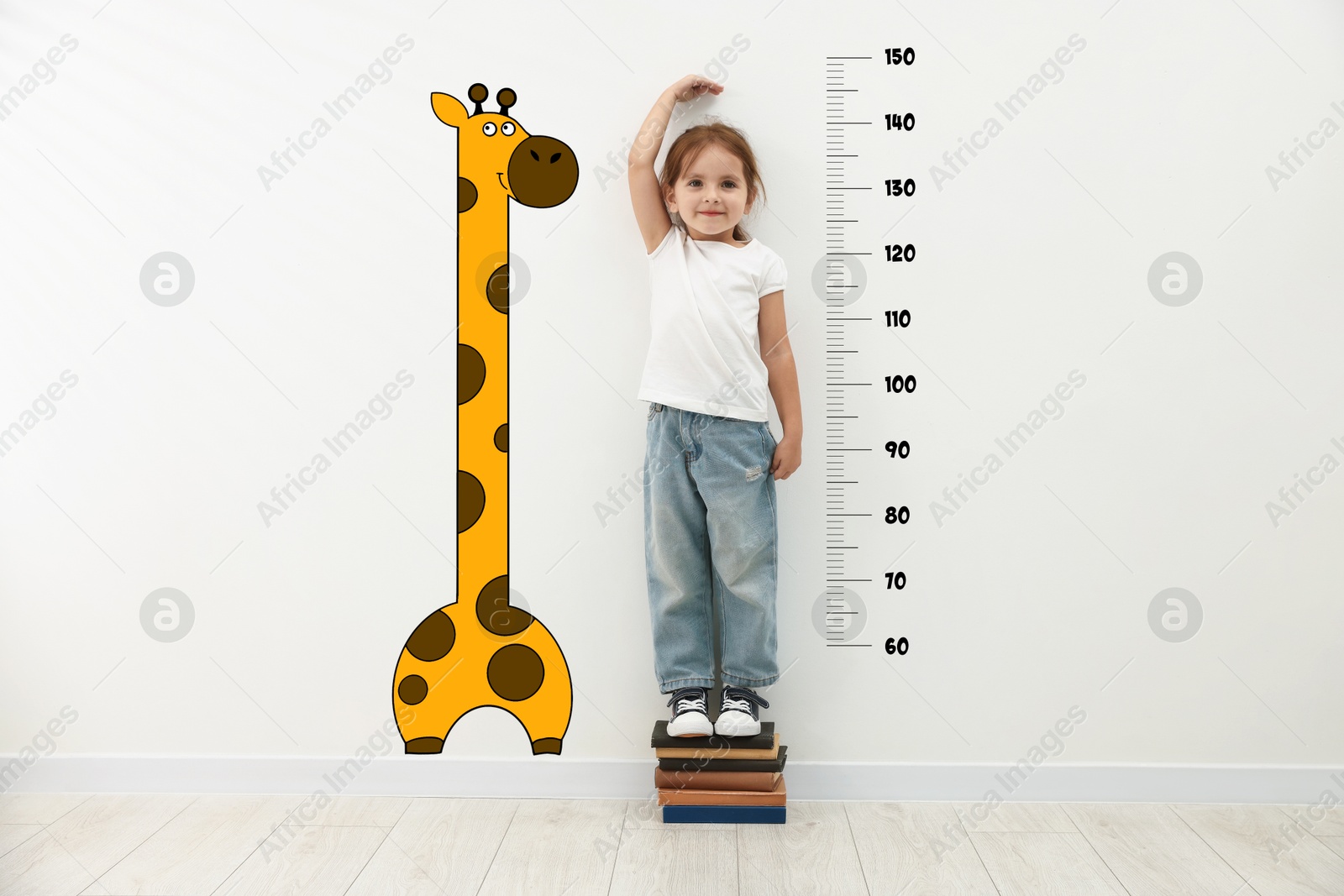 Image of Cute little girl standing on stacked books and measuring her height. White wall with scale and drawing of giraffe