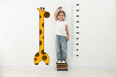 Image of Cute little girl standing on stacked books and measuring her height. White wall with scale and drawing of giraffe