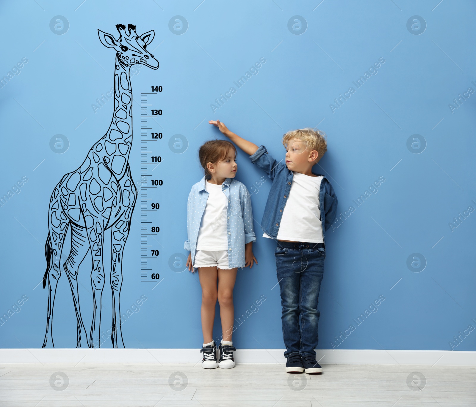 Image of Cute little children measuring their heights near blue wall with scale and drawing of giraffe